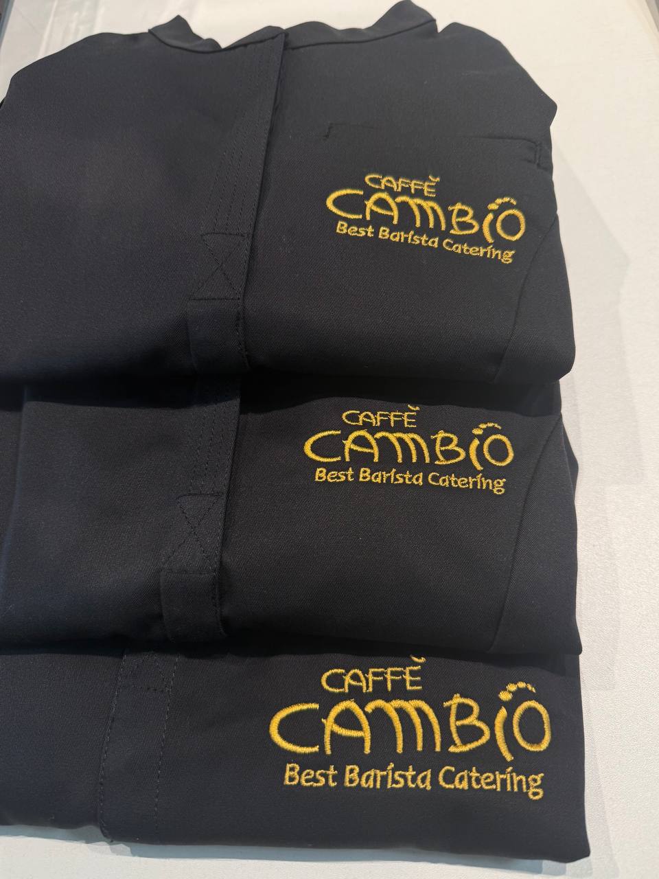 Gestapelte schwarze Poloshirts mit gelber Caffe Cambio Best Barista Catering Stickerei, Gastro-Teamwear Wien