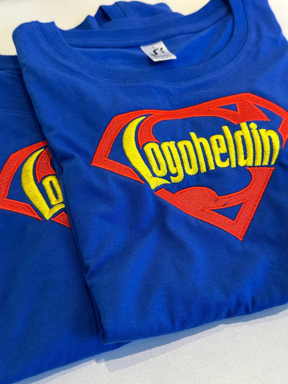 Blaues T-Shirt mit mehrfarbigem Logodruck, gefaltet auf hellem Untergrund