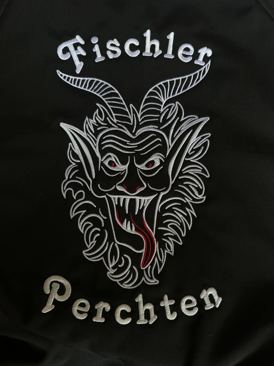 Weiß gesticktes Perchtenmotiv mit Schriftzug auf schwarzer Jacke