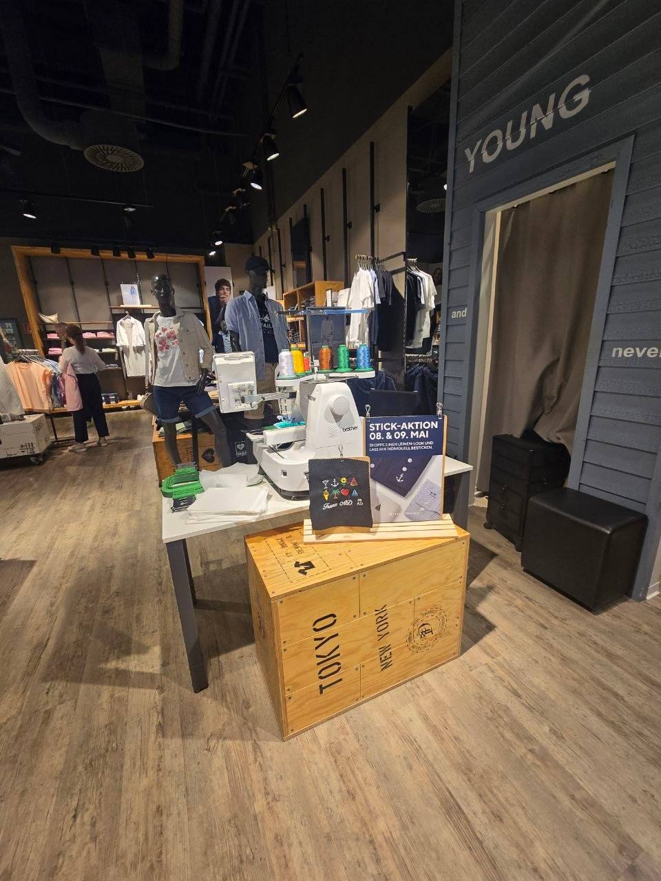Tom Tailor Eventstickerei Graz Setup im Store mit Brother Stickmaschine, Garnkonen und Tokyo New York Holzkiste vor der Young Section